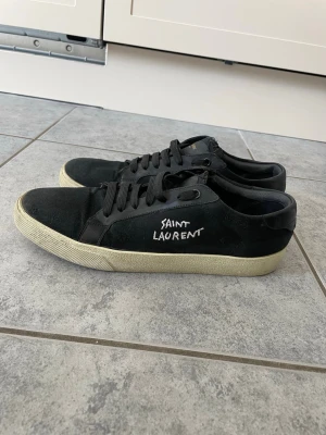 Svarta Saint Laurent sneakers - Svarta sneakers från Saint Laurent med vit logga på sidan. Skorna har låga skaft, svarta skosnören och vit sula. Materialet är en mix av canvas och läderdetaljer. Kommer med originalkartong och dustbag. Priset är inte hugget i sten, bara att hojta till för fler frågor!