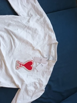 Vit sweatshirt från Ami med hjärtdetalj - Vit sweatshirt från Ami Alexandre Mattiussi med stor röd hjärtapplikation och bokstaven A på bröstet. Tröjan har rund halsringning och ribbade muddar. Perfekt för dig som gillar streetwear och snygga detaljer.