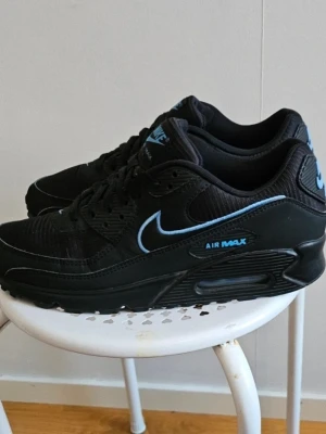 Nike Air Max 90 svart/blå sneakers - Svarta orginal Nike Air Max 90 sneakers med blå detaljer och logga. Klassisk siluett med mesh och läderpaneler, samt synlig Air Max-dämpning i sulan. Snygg kontrast mellan svart bas och blå swoosh samt text. Perfekta för dig som gillar streetwear och sportig stil.använda någon enstaka gång så som helt nya 