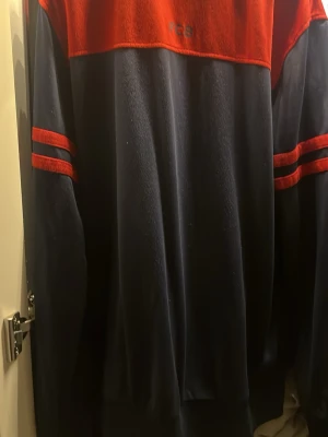 Barcelona FCB sweatshirt marin/röd - Säljer en snygg sweatshirt från FC Barcelona i marinblått och rött med klubbens broderade emblem på bröstet. Tröjan har röda ränder på ärmarna och är tillverkad i mjukt material, perfekt för dig som älskar fotboll och vill representera ditt lag. 