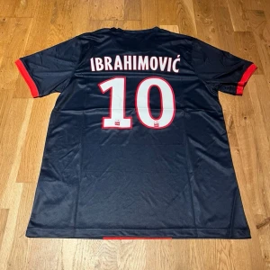 PSG Ibrahimović matchtröja - Mörkblå PSG matchtröja från Nike med röd vertikal rand och röda detaljer vid ärmslut. På ryggen står det 'IBRAHIMOVIĆ' och nummer 10. Framtill finns PSG-logga och Fly Emirates-tryck. Tillverkad i Dri-FIT material för bra ventilation.