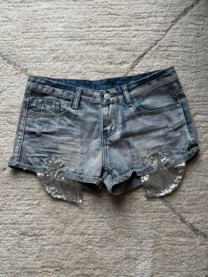 Ljusblå jeansshorts - Ljusblå lågmidjade jeansshorts med paljettdetaljer, en liten fläck framtill som knappt märks💕Passar storlek S och M