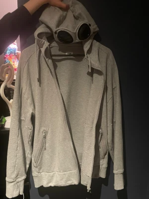 Grå hoodie med goggles från C.P. Company - Säljer en grå hoodie från C.P. Company med dragkedja, två fickor med dragkedja och ribbade muddar. Hoodien har en unik huva med inbyggda goggles och justerbara snören. Perfekt för dig som vill sticka ut med streetwear-stil.