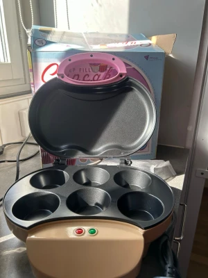 Cupcake Maker rosa för 6 muffins - Rosa elektrisk Cupcake Maker med plats för 6 muffins. Maskinen har rund form, lock med cupcake-tryck och indikatorlampor framtill. Tillverkad i metall och plast, perfekt för att baka cupcakes snabbt och enkelt hemma.