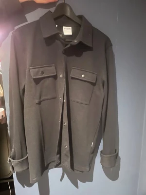 Svart overshirt från selected home  - Stilren svart overshirt från selected home med klassisk krage, två stora bröstfickor med lock och knappar samt knäppning framtill. Overshirten har långa ärmar med uppvikta muddar och är tillverkad i ett mjukt material som ger en avslappnad look.