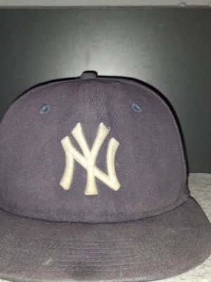 Mörkblå New Era Yankees keps - Säljer en mörkblå keps från New Era med klassiskt New York Yankees broderat vitt logotyp framtill. Kepsen har rak skärm och ventilerande hål upptill. Materialet är bomullstyg och loggan på sidan visar New Era-märket.
