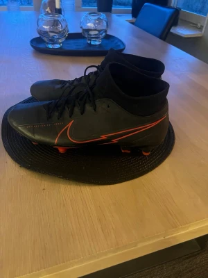 Nike svarta fotbollsskor med röd detalj - Säljer ett par svarta Nike fotbollsskor med röda detaljer och klassisk Swoosh-logga på sidan. Skorna har snörning, högre strumpliknande skaft och dobbar för bra grepp på planen. Perfekta för dig som vill ha stilrena och sportiga skor.