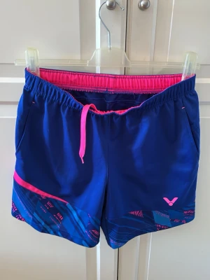 Blå sportshorts från Victor - Snygga blå sportshorts från Victor med rosa detaljer och mönstrad nederkant. Shortsen har elastisk midja med rosa snörning. Tillverkade i ett lätt och snabbtorkande syntetmaterial. Storlek M.  I fint skick, förutom en liten tråddraging (se bild).