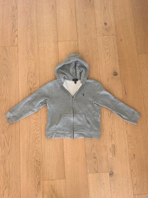 Grå Ralph Lauren hoodie med zip - En grå Ralph Lauren hoodie med zip, fint skick. Storlek 14-16 år enligt Ralph Lauren, tycker att den är liten i storlek. Kontakta vid minsta fundering! 