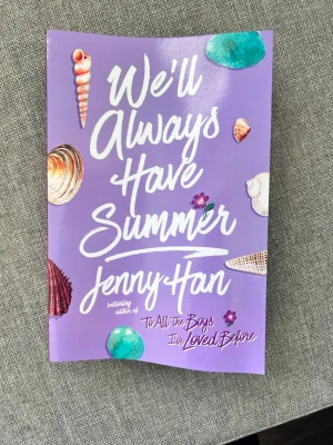 We’ll Always Have Summer av Jenny Han - En färgstark pocketbok med lila omslag och strandtema, dekorerad med snäckor och blommor. Boken är skriven av Jenny Han och är en del av en populär trilogi. Perfekt för dig som gillar romantisk ungdomslitteratur och sommarvibbar.