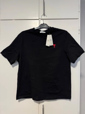 Svart t-shirt från Ami Paris - Svart t-shirt från Ami Paris med klassisk passform och korta ärmar. På bröstet finns ett litet rött hjärta med A-logga. Tillverkad i mjuk bomull, perfekt för en stilren och enkel look. Ny oandvänd.