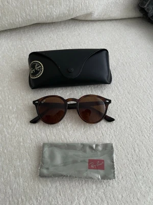 Bruna Ray-Ban solglasögon runda - Säljer ett par snygga runda solglasögon från Ray-Ban i brunsköldpaddsfärgad plast med mörka glas. Klassisk modell med Ray-Ban-logga på linsen och skalmen. Fodral och putsduk från Ray-Ban ingår.