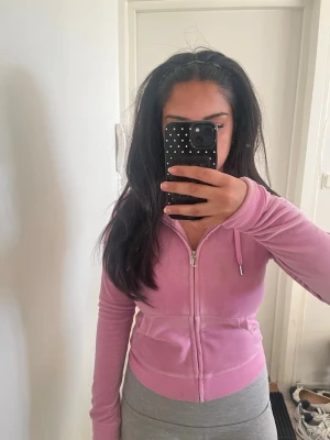 Rosa kofta  från Juicy Couture - Säljer en rosa velourhoodie från Juicy Couture med dragkedja framtill, huva och fickor. Hoodien har ett litet Juicy Couture-logotypbroderi på bröstet och ribbade muddar. Perfekt för en chill och trendig look.
