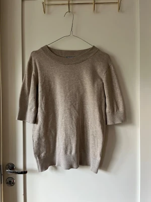 Beige stickad tröja  - En enkel och stilren beige stickad tröja med halvlånga ärmar och bred ribbad rundhals. Tröjan har en rak passform och är tillverkad i ett mjukt, tunt stickat material som passar perfekt till lager på lager. Perfekt basplagg för din garderob.