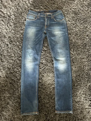 Blå Nudie Jeans Grim Tim - Säljer ett par klassiska blå jeans från Nudie Jeans med raka ben och snygg slitning framtill. Jeansen har fem fickor, orange sömmar och Nudie-logga på bakfickan. Tillverkade i bomull med en skön passform och tidlös look. 30/32. Riktigt bra skick med en liten lagning som iprincip inte syns. Riktig fet wash. Pris kan diskuteras. 