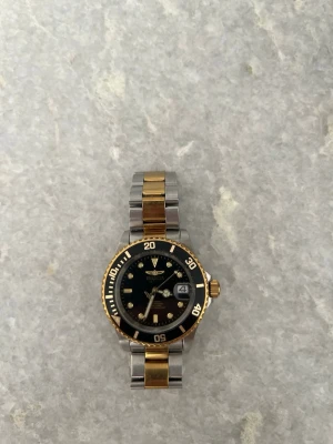 Invicta klocka  - Stilren Invicta klocka med svart urtavla, datumvisning och tvåfärgad länk i guld och silver. Klockan har tydliga indexmarkeringar och en klassisk rund form. Perfekt accessoar för dig som gillar en lyxig och tidlös look.
