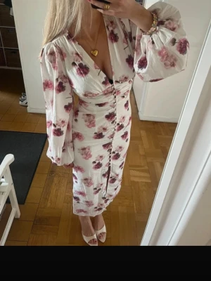 Vit blommig långklänning med puffärm - Romantisk långklänning i vit bomull med rosa och lila blommigt mönster. Klänningen har djup v-ringning, knappar framtill och långa puffärmar. Perfekt för sommarens alla tillfällen och ger en feminin och trendig look.