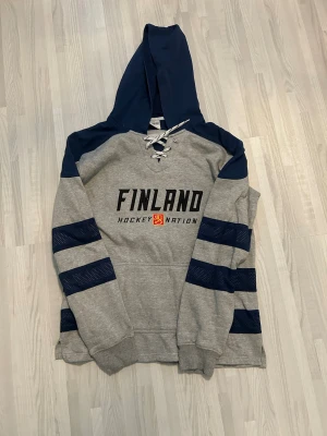 Grå Finland Hockey Nation hoodie - Snygg grå hoodie med mörkblå detaljer och huva. På bröstet står det 'Finland Hockey Nation' med ett broderat lejonmärke. Hoodien har snörning vid halsen och mörkblå ränder på ärmarna. Perfekt för dig som gillar hockey och vill visa ditt stöd för Finland.