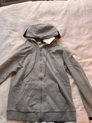 Grå Moncler hoodie med dragkedja - Snygg grå hoodie från Moncler med dragkedja framtill, huva med dragsko och klassisk logga på ärmen. Mjuk bomullskänsla och känguruficka framtill. Perfekt för en chill och stilren look. Den har ett litet håll vid fickan man kan fixas enkelt 
