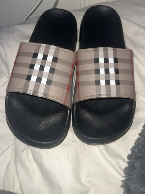 Burberry slides med rutigt mönster - Snygga slides från Burberry med klassiskt beige, svart, vitt och rött rutmönster över foten. Sulan är svart och har en bekväm formad design. Perfekta för chill dagar eller när du vill ha en stilren look med streetkänsla.
