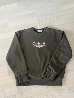 Olivgrön Carhartt sweatshirt - Olivgrön sweatshirt från Carhartt med broderad logga och texten 'work in progress' på bröstet. Tröjan har rund halsringning, ribbade muddar och är tillverkad i mjuk bomullsblandning. Perfekt för en avslappnad streetwear-look.
