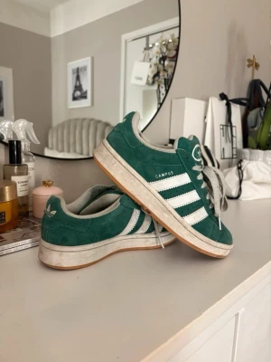 Adidas Campus gröna sneakers - stl 37 - Gröna Adidas Campus sneakers i mocka med klassiska vita ränder på sidorna och vit logga på plösen. Skorna har lågt skaft, vita skosnören och gummisula i ljusbrunt. Perfekta för en avslappnad och trendig stil.