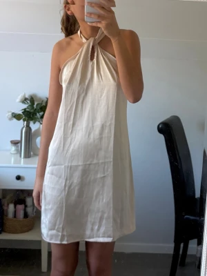 Vit halterneck klänning i satin - Snygg vit kortklänning med halterneck och knyt i nacken från Shein. Klänningen har en avslappnad passform och är tillverkad i svalt linne, perfekt för varma dagar. Enkel och stilren design som ger en fräsch look. Aldrig använd!