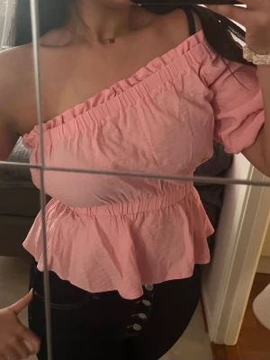 Rosa omlott blus med oneshoulder - Supersöt rosa blus med volangkant och resår i midjan. Toppen har en ärm och en axel är bar för en trendig oneshoulder-look. 💘💘