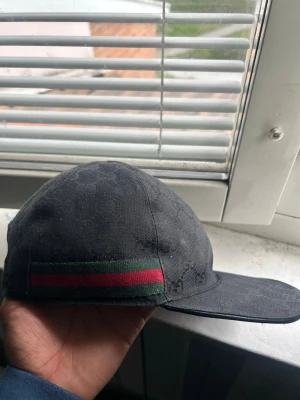 Gucci keps svart  - Svart keps från Gucci med diskret GG-monogram över hela tyget och klassiska gröna och röda ränder på sidan. Kepsen är i bomull och har en böjd skärm. Storlek S, 57 cm. Perfekt för dig som vill ha en stilren och lyxig accessoar.