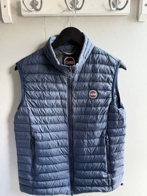 Blå dunväst från Colmar - Snygg blå dunväst från Colmar med quiltad design och hög krage. Västen har två dragkedjefickor framtill och en tydlig Colmar-logga på bröstet. Perfekt för lager-på-lager och enkel att matcha med olika outfits.