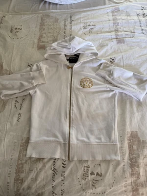 Vit hoodie från Versace Jeans - Snygg vit hoodie från Versace Jeans med dragkedja och guldfärgad logga på bröstet. Perfekt för en stilren och trendig look. Den är lite gul vid luvan pågrund av jag använt parfym som har fått vätskan att fastna. Men de går att tvätta bort med rätt material, ny pris går på 3000, Storlek är mellan S-M det står inget på tröjan 