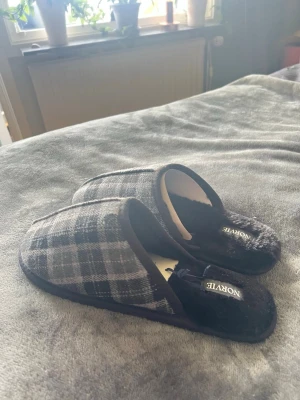Rutiga tofflor från Norvite - Mysiga tofflor från Norvite i svart och grått rutigt tyg med mjuk, fluffig insida. Perfekta att glida runt i hemma när du vill ha det extra bekvämt. Klassisk slip-in modell med låg häl och textilmaterial. Storleken är både 42/43