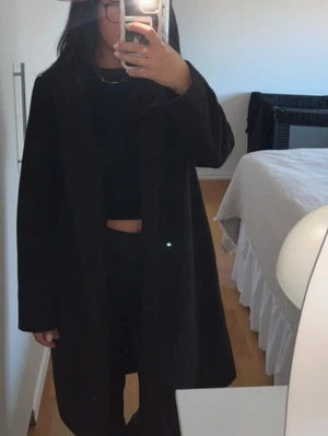 Svart kappa från Zara - Stilren svart kappa från Zara med rak passform och klassisk krage. Kappan är lång och har en enkel, tidlös design utan synliga knappar. Perfekt för dig som gillar minimalistisk stil och vill ha en snygg jacka till hösten eller vintern.
