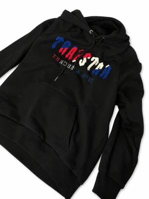 Svart Trapstar hoodie med brodyr - Svart hoodie från Trapstar med broderad logga i blått, rött och vitt framtill. Klassisk känguruficka och justerbar huva med dragsko. Mjuk bomullskänsla och avslappnad passform, perfekt för en streetwear-look. Lite knopprig inuti och lite utanpå men går såklart att åtgärda- scrolla till andra bilden för egentagen bild