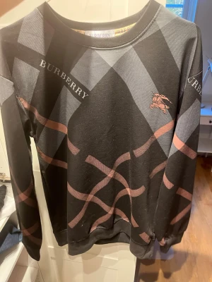 Svart Burberry sweatshirt med mönster - Snygg svart sweatshirt från Burberry med klassiskt rutmönster i grått och brunt samt logga på bröstet. Tröjan har rund halsringning och långa ärmar. Materialet känns mjukt och bekvämt, perfekt för en trendig och avslappnad stil.