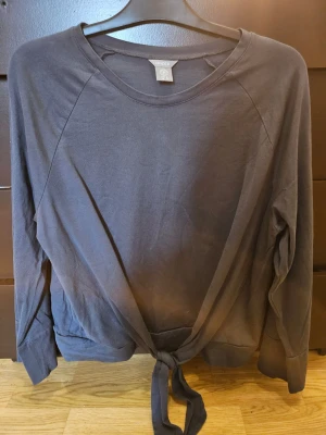 Mörkgrå långärmad topp från Lindex - Snygg mörkgrå långärmad tröja från Lindex med rund halsringning och knyt framtill. Tillverkad i mjuk bomull som känns skön mot huden. Perfekt att matcha med jeans eller kjol för en avslappnad look.
