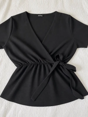 Svart omlottblus från Boohoo - Stilren svart omlottblus från Boohoo med knytband i midjan och v-ringad hals. Blusen har långa, lätt utsvängda ärmar och ett mjukt, tunt tyg som ger ett snyggt fall. Perfekt att matcha med jeans eller kjol för en trendig look.