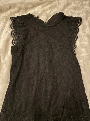 Svart spetsblus med volangärm - Snygg svart blus i spets med detaljerade volangärmar och rund halsringning. Blusen är ärmlös och har ett litet knapphål bak i nacken. Perfekt för dig som gillar feminina och stilrena plagg.