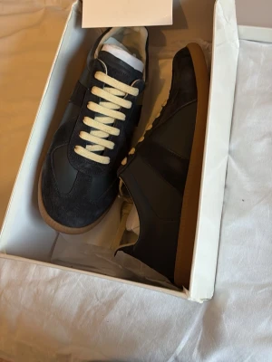 Maison Margiela Gats - Snygga svarta sneakers med beige sula och kontrasterande beigea skosnören. Ovandelen är i en mix av mocka och slätt läder för en stilren look. Klassisk låg modell som passar till det mesta. Perfekta för dig som gillar minimalistisk och tidlös design. 