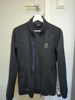 Svart softshell från Haglöfs -  från Haglöfs med dragkedja framtill och två sidofickor med dragkedja. Jackan har en hög krage och diskret logga på bröstet. Tillverkad i ett lätt och smidigt material som skyddar mot vind. Perfekt för utomhusaktiviteter.