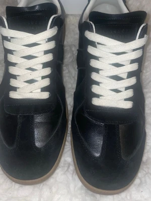 Masion margiela  - Masion margiela Stilrena svarta sneakers med vit insida och kontrasterande vita skosnören. Ovandelen är i en mix av läder och mocka, och sulan är i gummi med en klassisk brun färg. Perfekta för en clean och tidlös look.