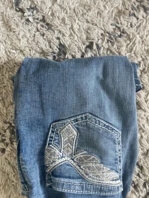 Blå slitna jeans med broderad ficka - Säljer ett par ljusblå jeans med slitningar på benen och en stor broderad detalj på bakfickan. Jeansen har en klassisk femficksmodell och är gjorda i denim. Perfekta för dig som gillar en avslappnad och trendig stil.