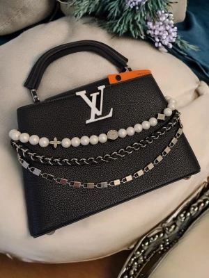 Iouis Vuitton Capucines - Design: En del av den ikoniska Capucines-linjen, namngiven efter Iouis Vuittons första butik i Paris (öppnad 1854).Den är djupt svart med en tydlig textur i lädret, vilket ger ett sofistikerat och robust intryck. Väskan utmärker sig genom sin eleganta dekorationskedja som hänger i en båge över framsidan; kedjan består av tre rader med stora vita pärlor, en klassisk metallkedja och en modernare länk med fyrkantiga element.