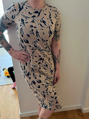 Beige mönstrad kort klänning - Säljer en beige kortklänning med svart och mörkblått abstrakt mönster. Klänningen har rund hals, korta ärmar och en figurnära passform. Perfekt för dig som gillar en stilren men ändå unik look.