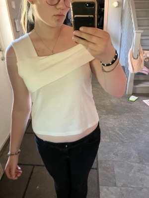 Vit offshoulder topp med asymmetrisk kant - Snygg vit topp med offshoulder-design och asymmetrisk kant upptill. Toppen är ärmlös och har en stilren, modern look som passar perfekt till jeans eller kjol. Materialet känns mjukt och stretchigt, vilket gör den bekväm att bära hela dagen.