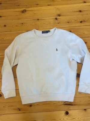 Beige sweatshirt från Polo Ralph Lauren - Klassisk beige sweatshirt från Polo Ralph Lauren med rund halsringning och diskret broderad logga på bröstet. Tröjan har ribbade muddar vid ärmslut och nederkant, samt en relaxed passform. Perfekt för en clean och stilren look.