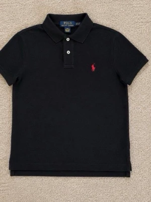 Svart pikétröja från Polo Ralph Lauren - Klassisk svart pikétröja från Polo Ralph Lauren med röd broderad logga på bröstet. Tröjan har korta ärmar, krage och två knappar framtill. Tillverkad i bomull för en bekväm och stilren look.