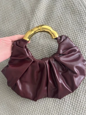 Vinröd handväska med guldfärgat handtag - Trendig vinröd handväska i mjukt, glansigt material med ett unikt, rundat handtag i guldfärgad metall. Väskan har en veckad design som ger en cool och modern look. Perfekt accessoar för att lyfta din outfit.