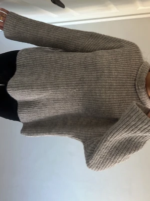 Grå ribbstickad tröja - Mysig grå stickad tröja med ribbstickat mönster och långa ärmar. Tröjan har en avslappnad passform och hög hals, perfekt för kyliga dagar. Passar till både jeans och kjol för en enkel och stilren look.
