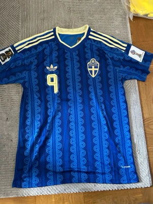 Blå Sverige fotbollströja Adidas - Säljer en blå fotbollströja från Adidas med Sveriges landslagsemblem och nummer 9 på bröstet. Tröjan har gula detaljer, klassiska Adidas-ränder på axlarna och ett retroinspirerat mönster. Perfekt för dig som vill stötta Sverige med stil.